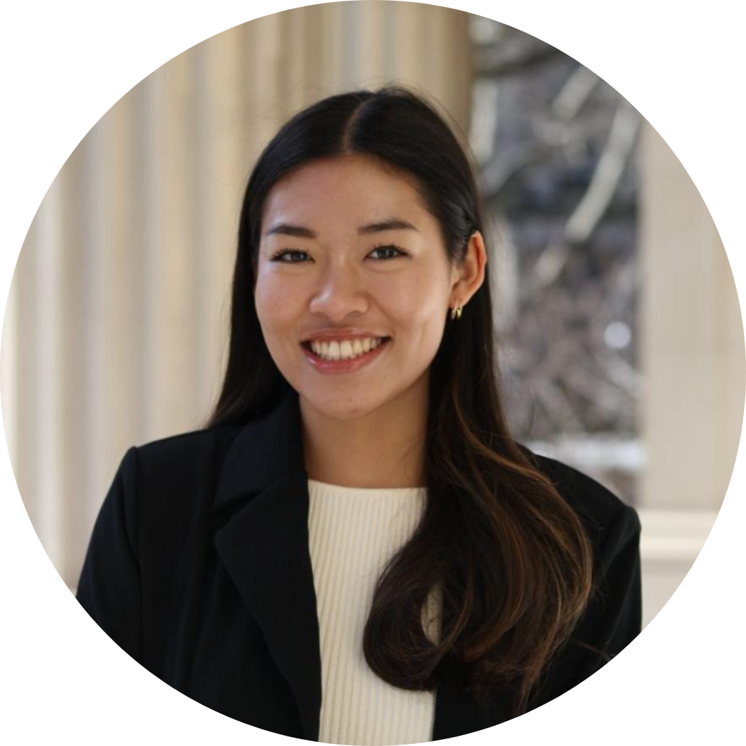 Allyce smiles and looks at the camera. She is wearing a black blazer, and white shirt, and stands in front of marble pillars.