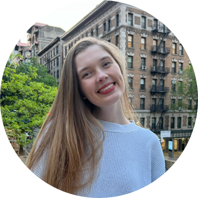 Hannah has long light brown hair and wears a blue sweater. She is smiling and looking at the camera. Hannah stands in front of trees and buildings.