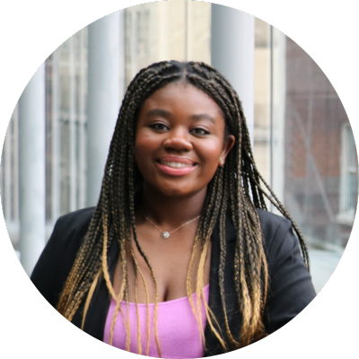 Sahmaya wears a pink top and black blazer and smiles with her mouth open. She is a Black woman with her hair in braids. She is posing inside in front of a background of glass with pillars.
